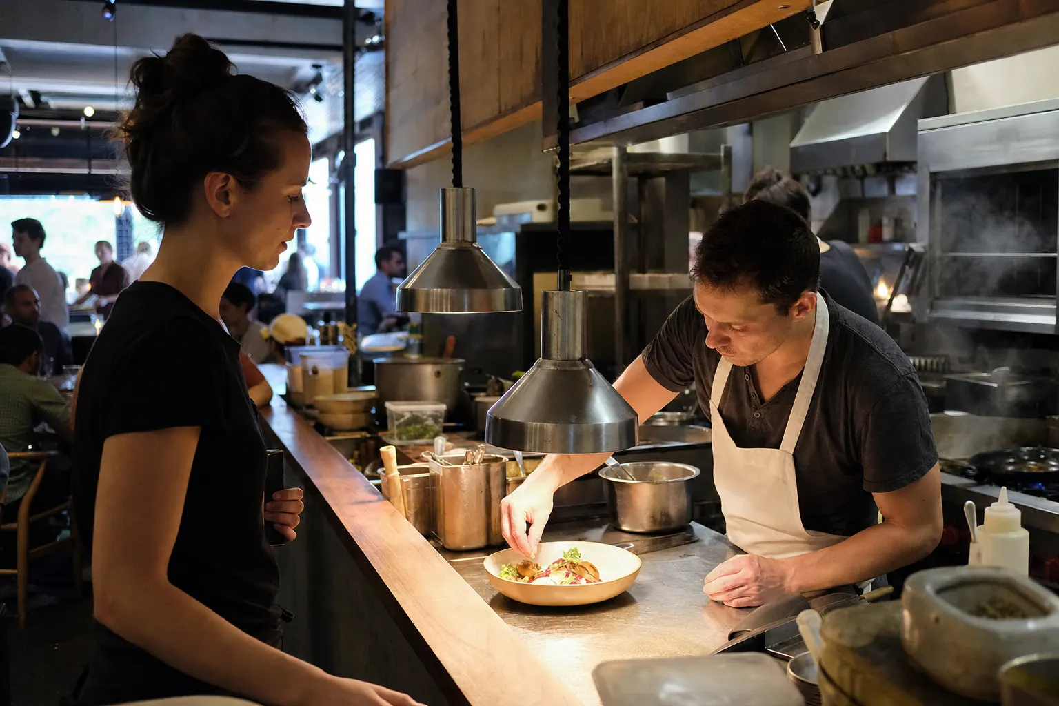 Line cook plating a dish under warm lights in a busy open restaurant kitchen